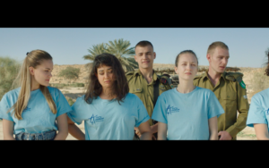 A group of young women in blue t shirts stand in front of two Israeli soldiers.