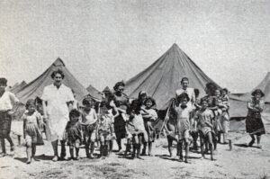 A group of children and adults stand in front of tents on sand