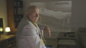 A bald man in a grey suit jacket turns around in his chair looking away from a projected image on a wall of a man covered in bandages in a bed.